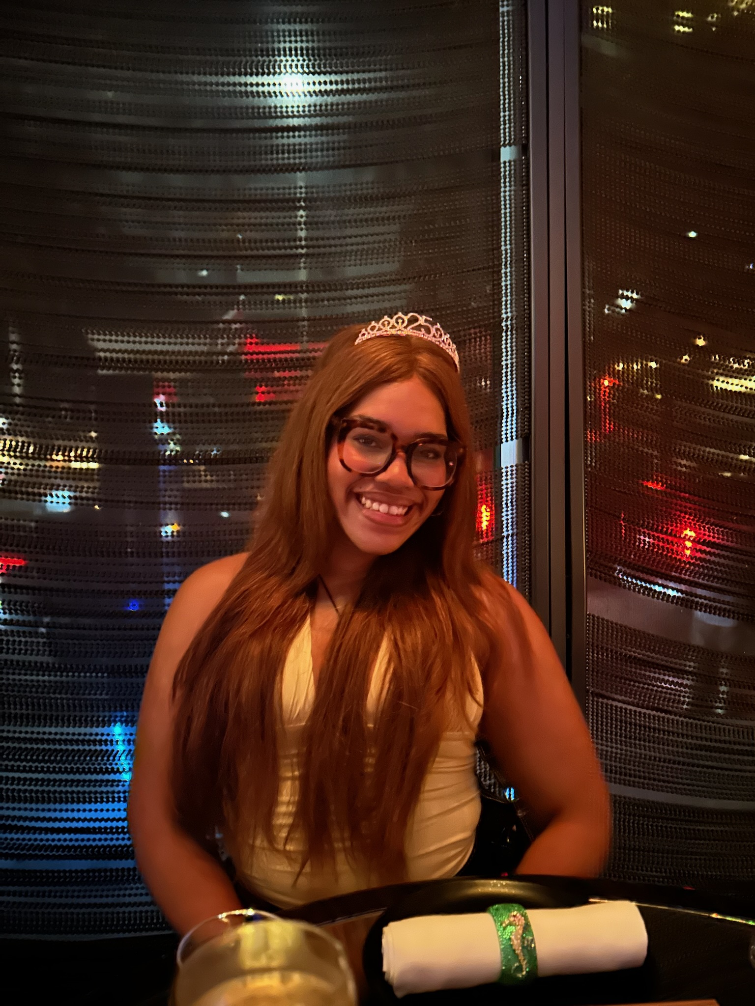 Jayda smiling in a tiara and glasses at a dinner table with city lights behind her